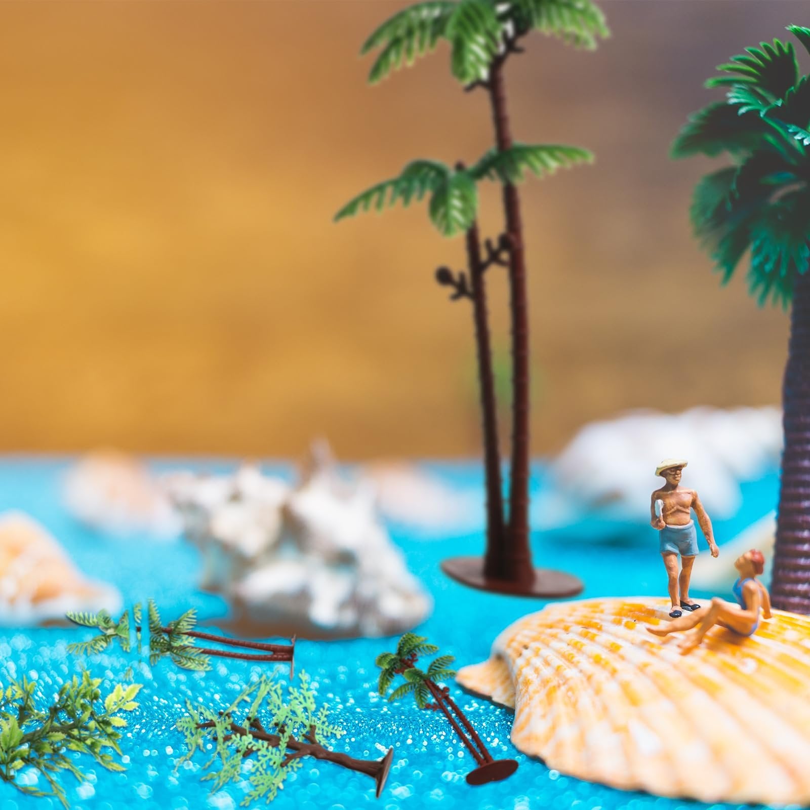 Miniature beach scene with two figures on a seashell, palm trees, and shells on blue glittery water.