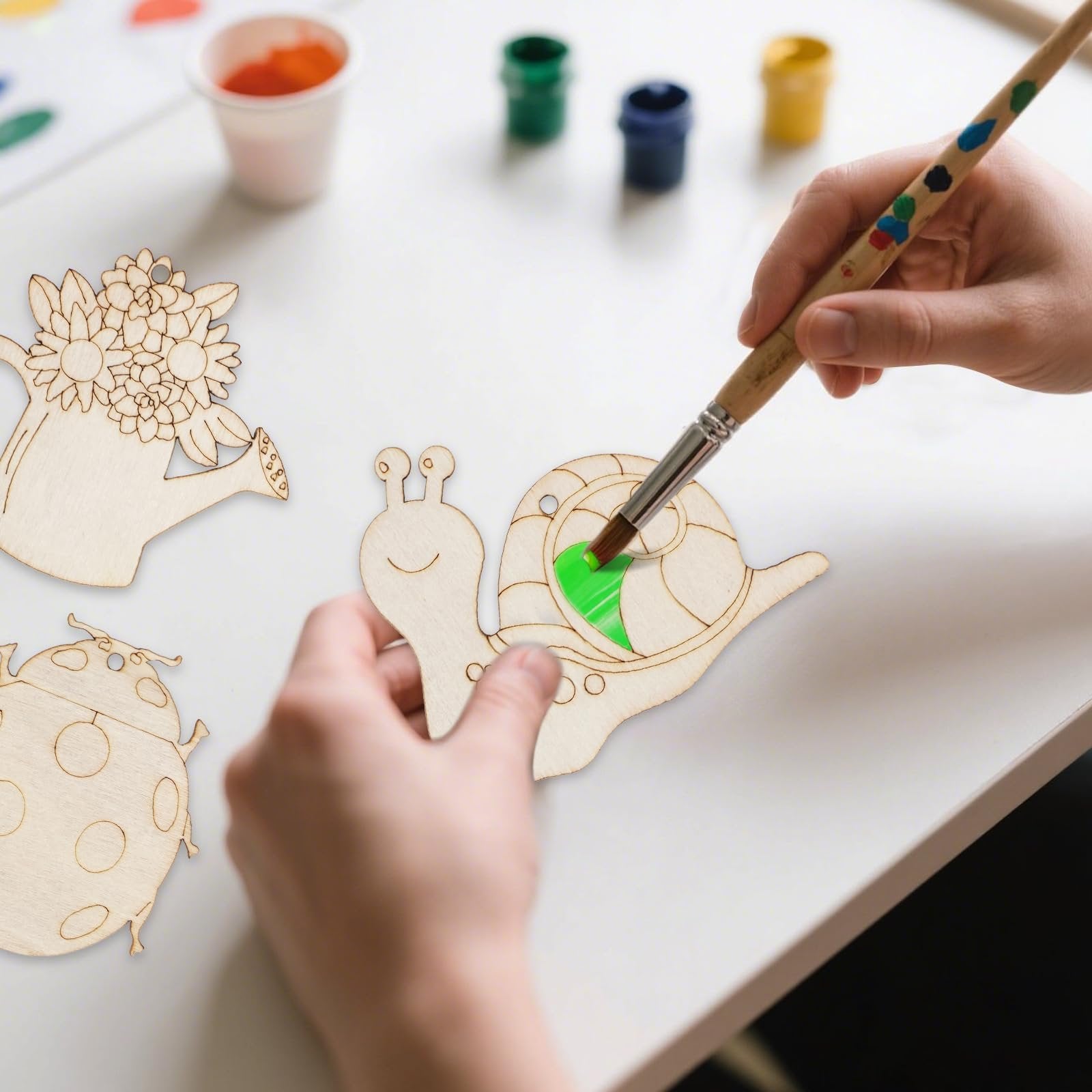 Hands painting unfinished wooden craft ornaments shaped like a ladybug, watering can with flowers, and a snail.