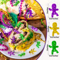 Mardi Gras king cake with purple, gold, and green plastic baby figures scattered on top and around it, with Mardi Gras beads and feathers.