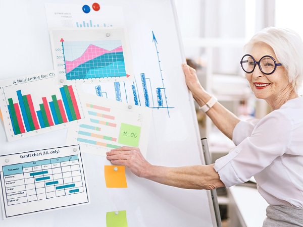 Elderly woman with glasses presenting charts and graphs on a whiteboard, using magnetic push pins.