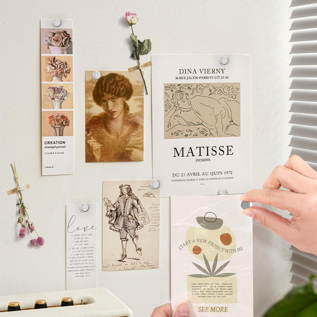 Silver magnetic push pins holding various art prints and photos on a wall, with a hand placing one pin.