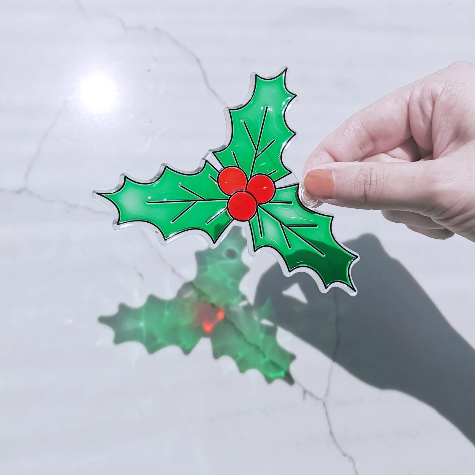 Hand holding a shiny green holly leaf suncatcher with red berries against a marble background