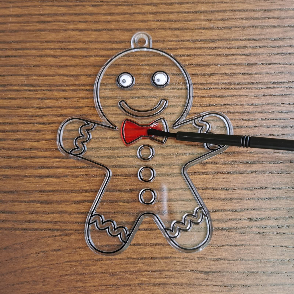 Clear gingerbread man suncatcher ornament with black outline, white eyes, red bow tie, and a brush applying paint to the bow tie, on a wooden background.