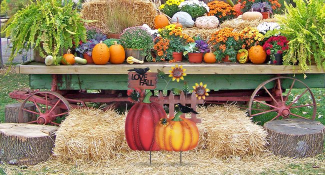 Decorative metal fall garden stakes with pumpkins, sunflowers, and crows, set against a backdrop of a rustic wagon filled with autumn plants and hay bales.