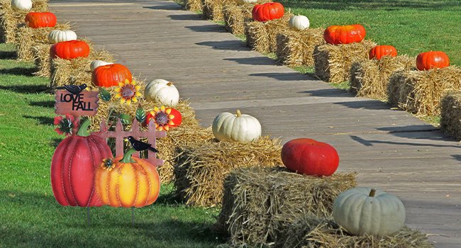Decorative metal garden stake with pumpkins, sunflowers, crows, and 'I LOVE FALL' sign, set along a path lined with hay bales and pumpkins.