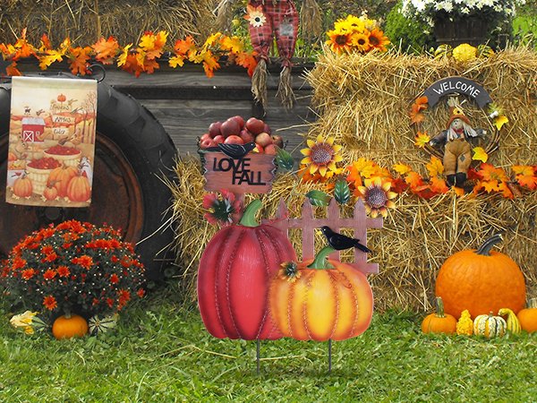 Decorative metal garden stakes with pumpkins, sunflowers, and a crow, set against hay bales and autumn foliage.