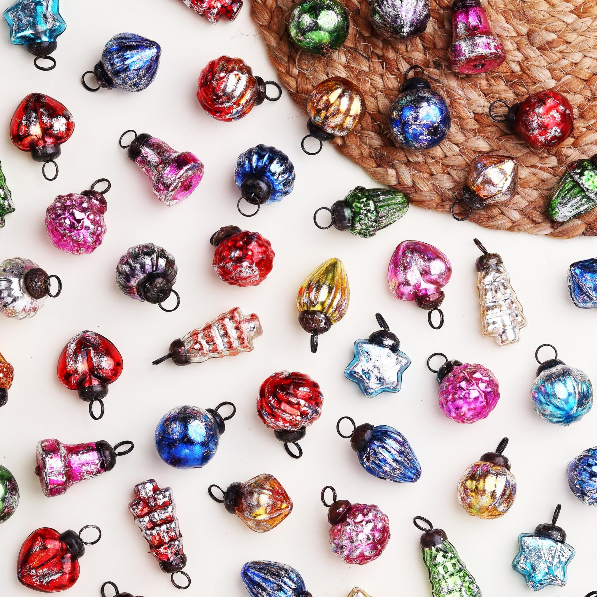 Assortment of 25 small, colorful, vintage-style glass Christmas ornaments scattered on a white surface with a woven mat.