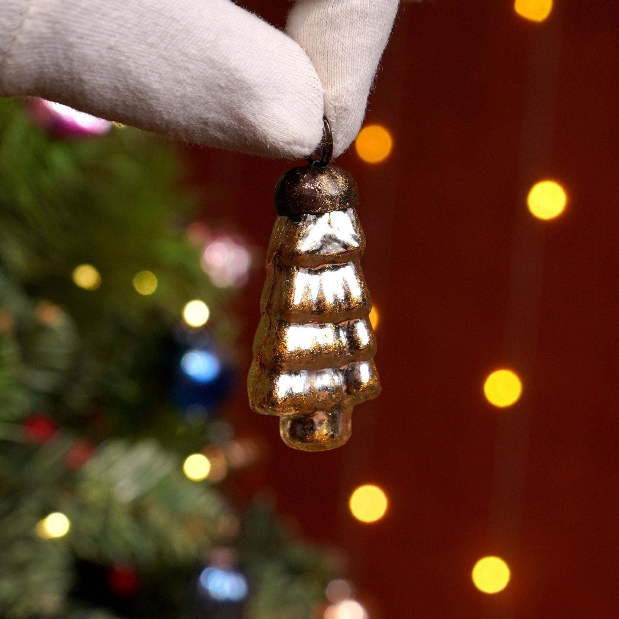 Hand wearing a white glove holds a small, golden, tree-shaped glass Christmas ornament against a blurred background of a decorated Christmas tree and bokeh lights.