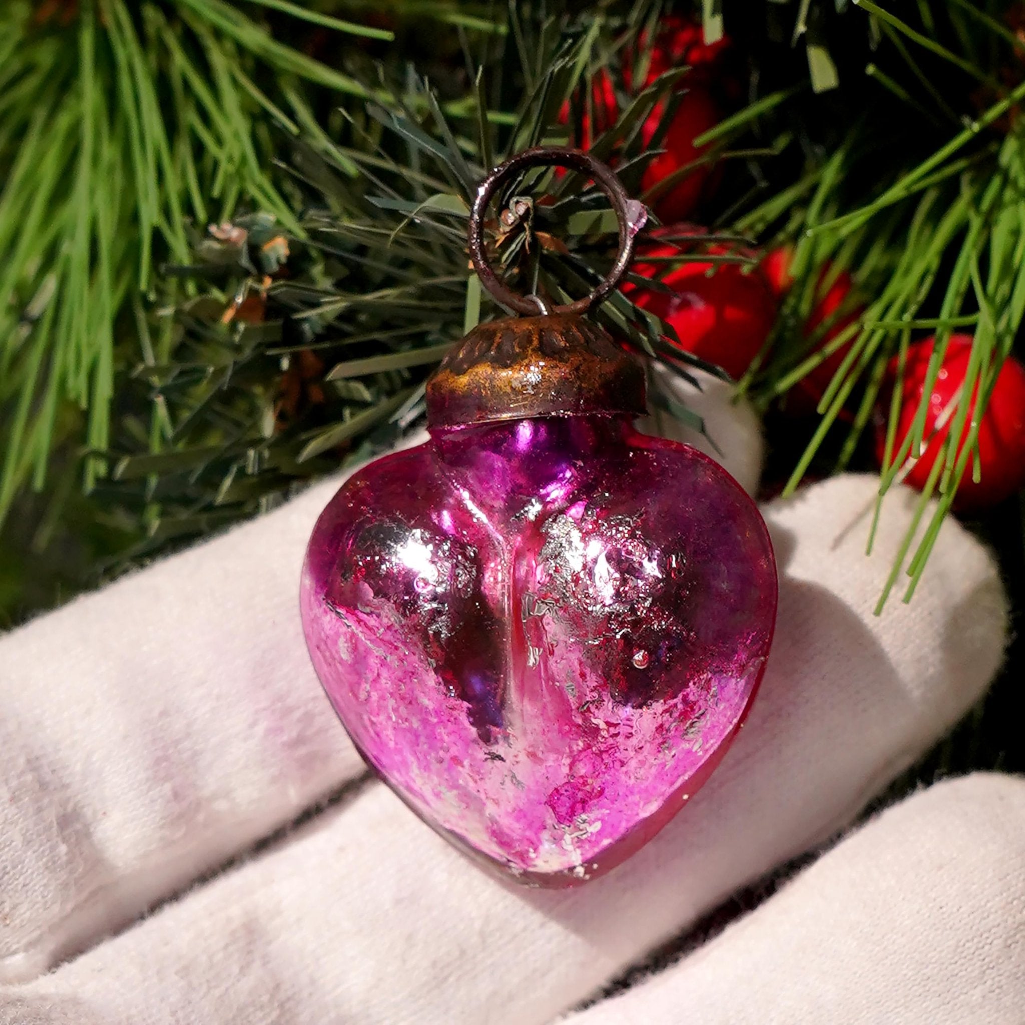 Close-up of a pink heart-shaped glass Christmas ornament held by a gloved hand, with pine branches and red berries in the background.