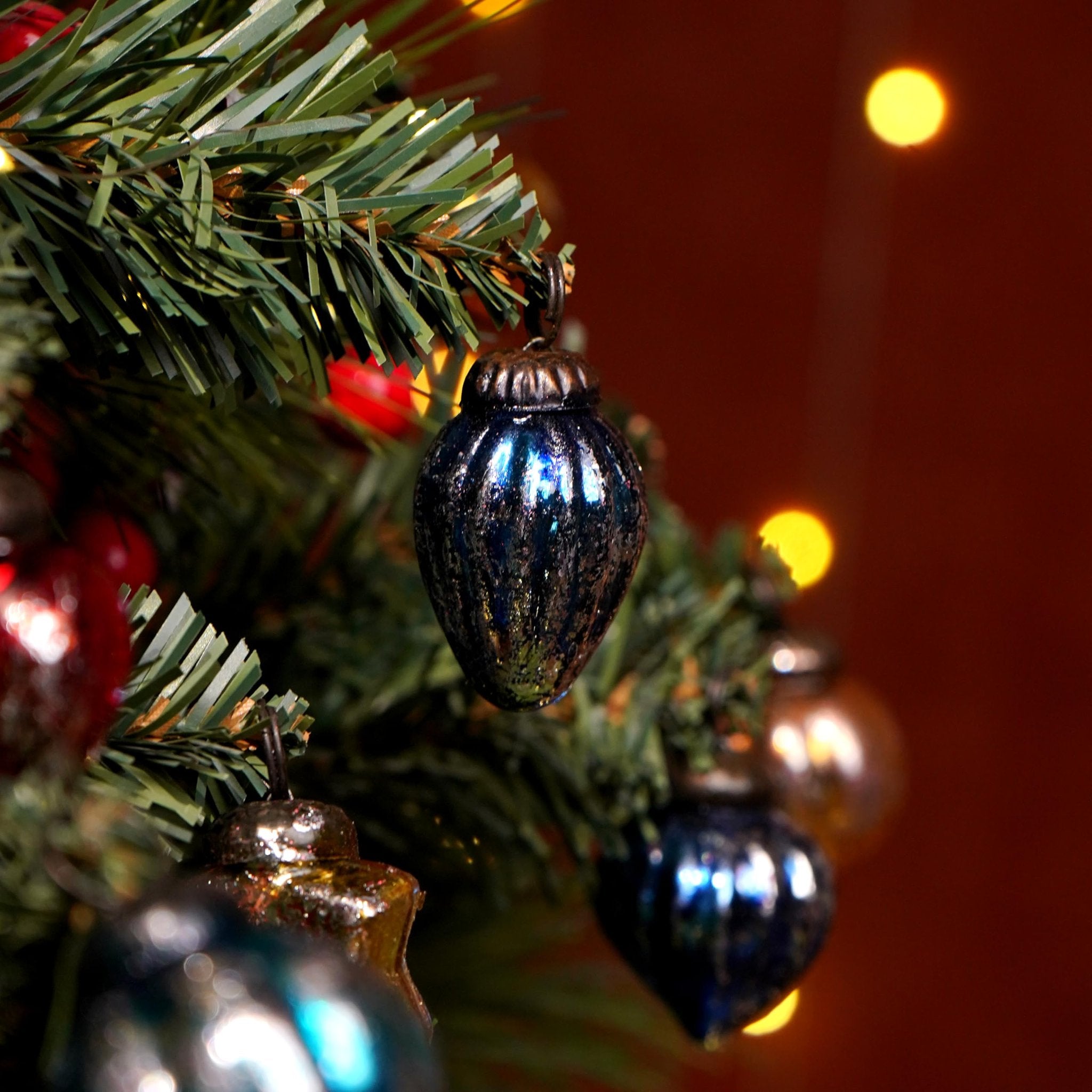 Close-up of vintage glass Christmas ornaments hanging on a pine tree branch with bokeh lights in the background.
