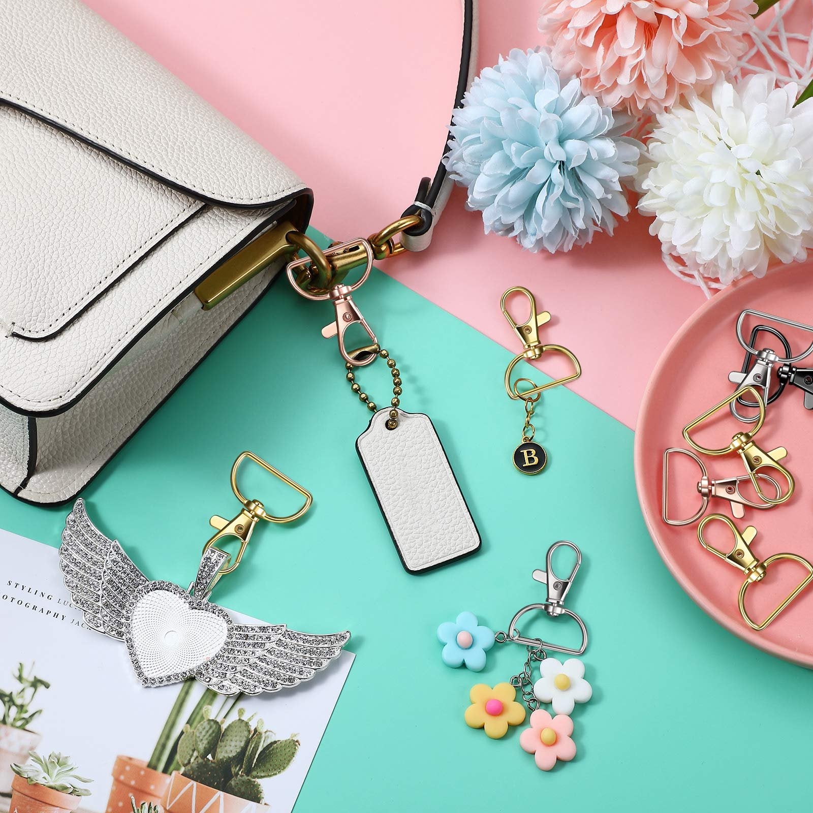 Keychain making kit components including swivel clasps, D-rings, a heart-shaped charm with wings, a blank tag, and flower charms, arranged around a white handbag.