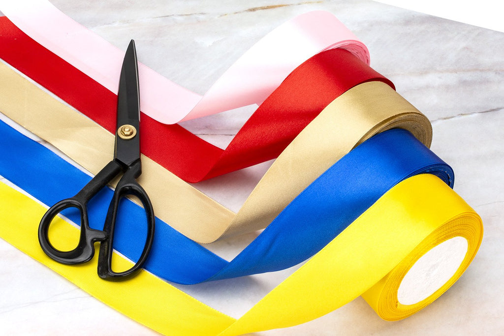 Rolls of satin ribbon in pink, red, gold, blue, and yellow with black scissors on a marble surface.