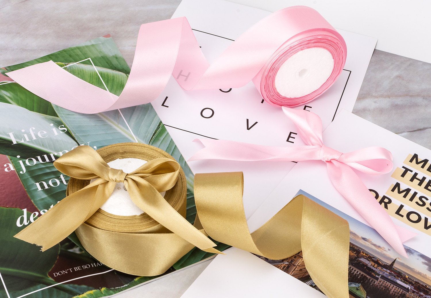 Two rolls of satin ribbon, one gold with a bow and one pink with a bow, on top of magazines and paper with text.