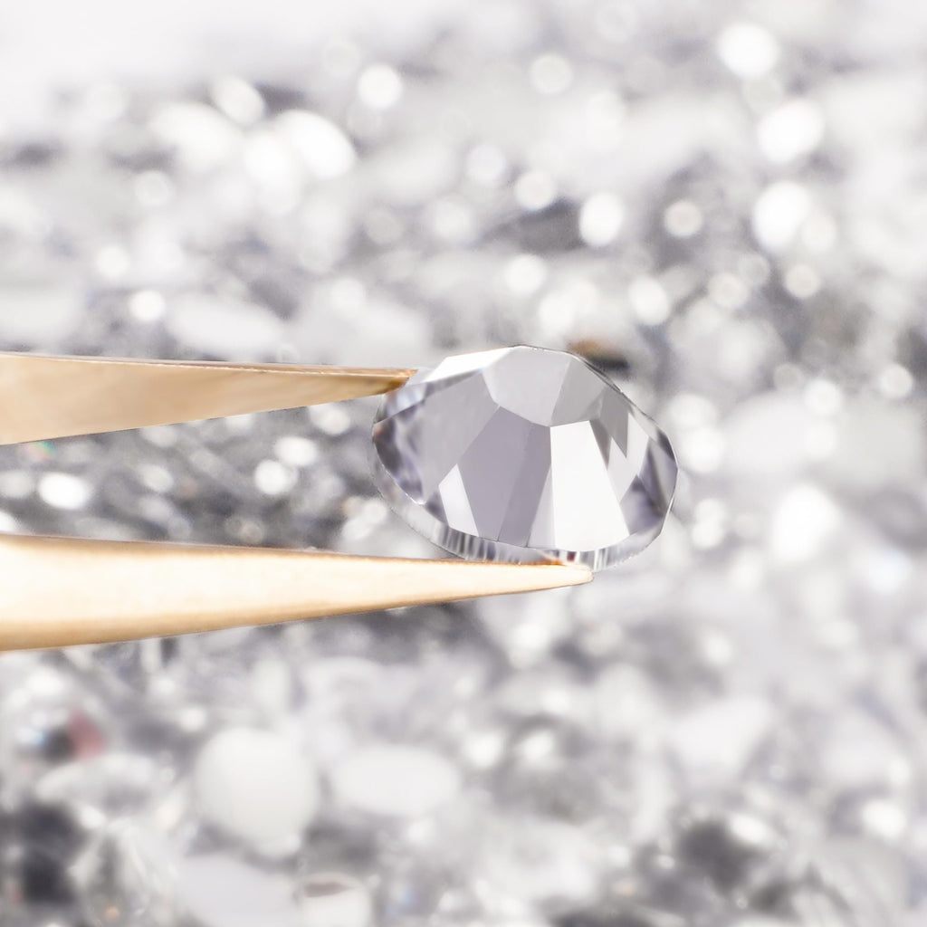 Close-up of a clear, faceted flatback rhinestone held by gold tweezers against a blurred background of many rhinestones.
