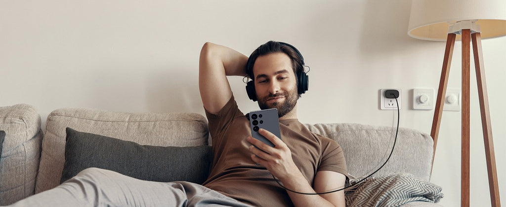 Man wearing headphones and holding a smartphone while relaxing on a couch, with a USB-C charger plugged into a wall outlet.