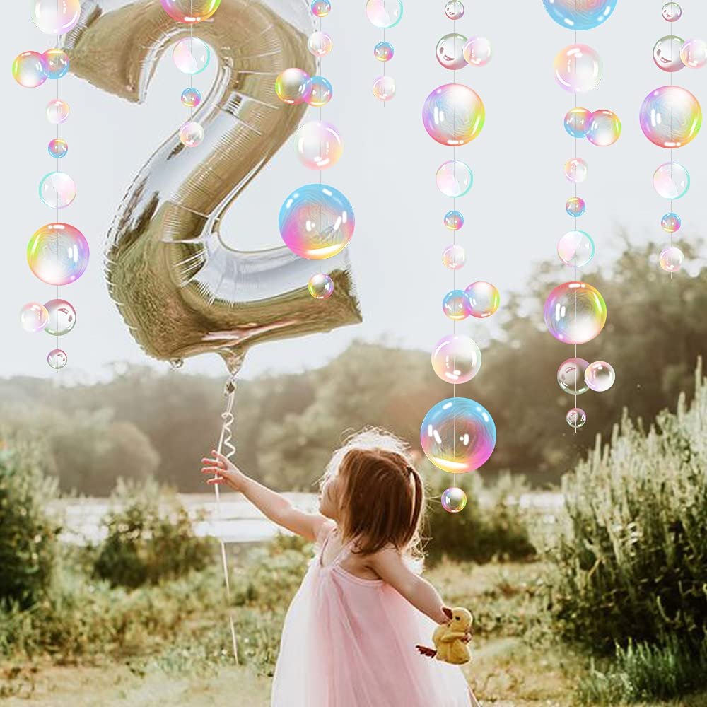 Young girl in pink dress reaching for a large silver number 2 balloon with iridescent bubble garlands hanging around her outdoors.