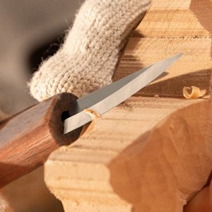 Close-up of a hand wearing a glove using a wood carving knife to shape a piece of wood.