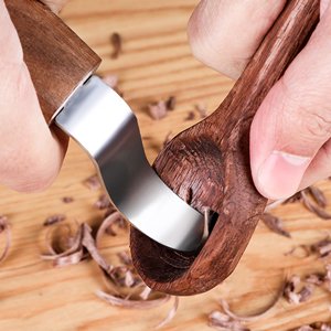 Hands carving a wooden spoon with a curved whittling knife, wood shavings scattered on a wooden surface.