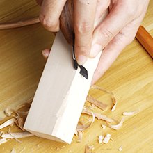 Close-up of hands carving a wooden block with a whittling knife, wood shavings scattered on a wooden surface.