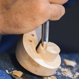 Close-up of a hand using a chisel knife to carve a wooden piece, showing wood shavings.