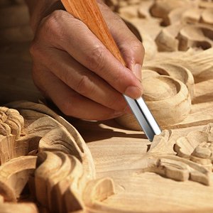 Close-up of a hand using a wood carving chisel to detail a wooden sculpture.