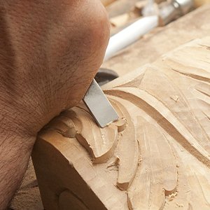 Close-up of a hand using a chisel knife to carve intricate details into a wooden sculpture.