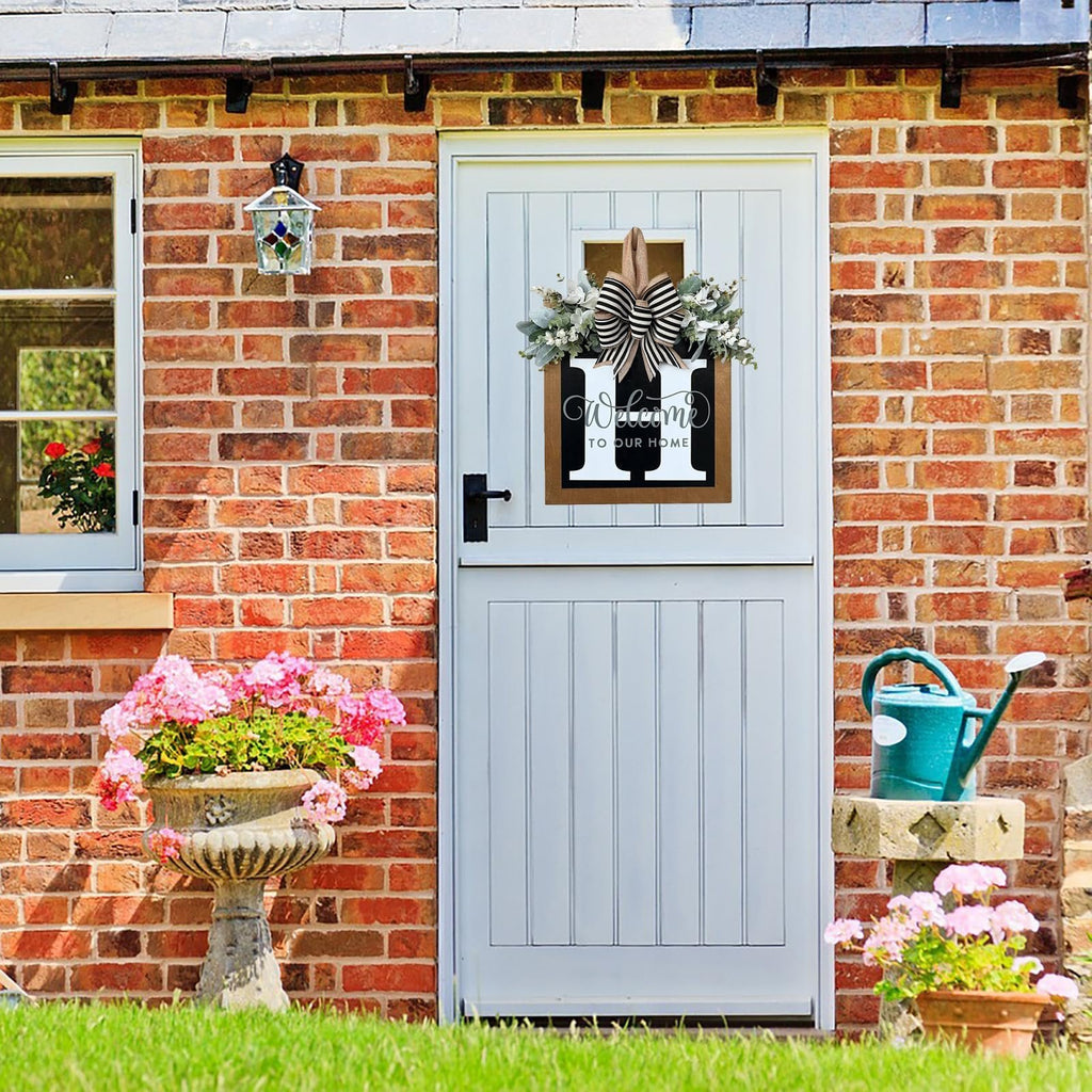 Front door decorated with a wreath featuring a large letter 'H', eucalyptus garland, and a striped bow. Text reads 'Welcome to Our Home'.