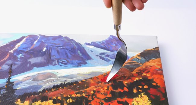 Artist using a palette knife to apply red paint to a landscape painting of mountains and trees