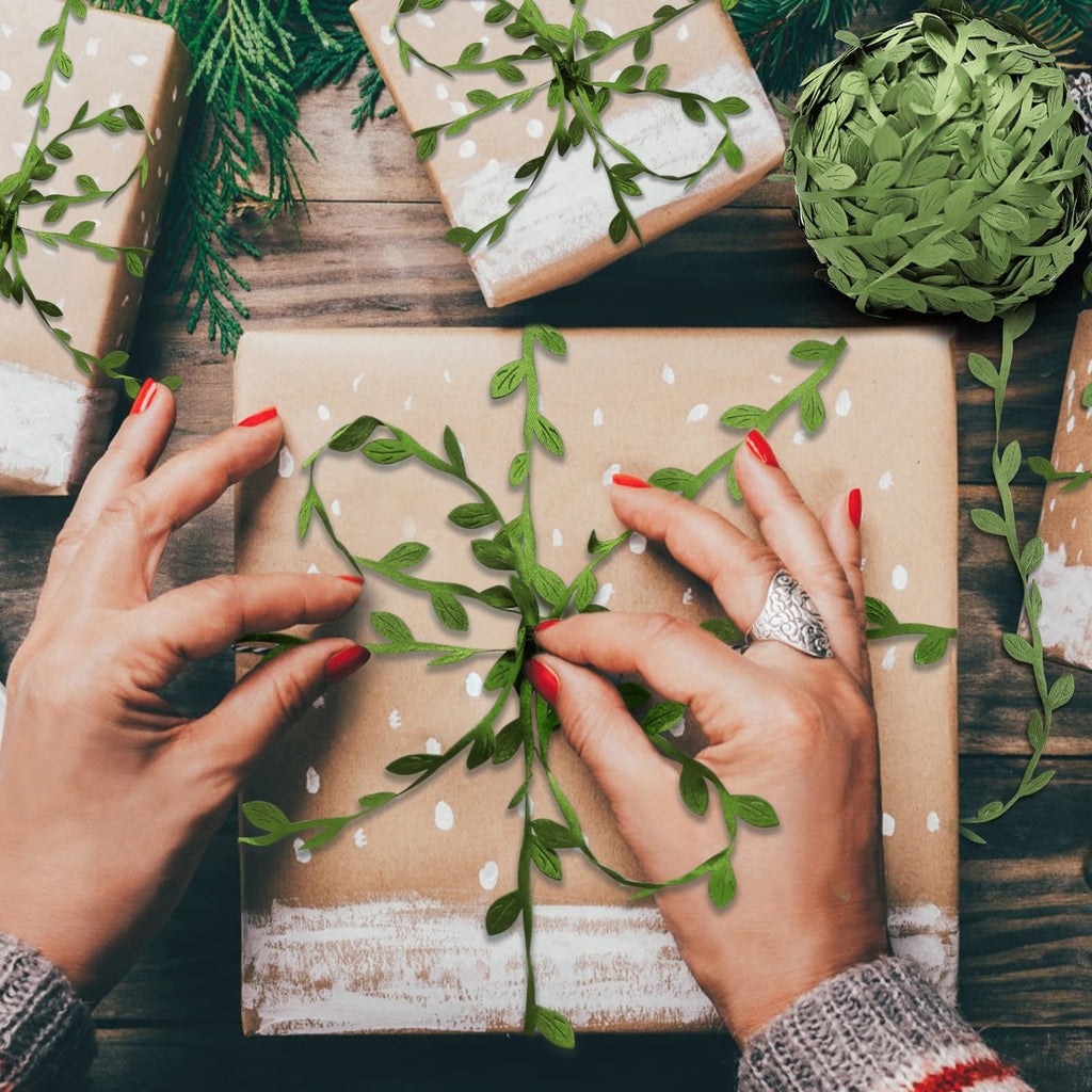 Hands decorating a gift box with artificial leaf vine ribbon, surrounded by other wrapped gifts and greenery.