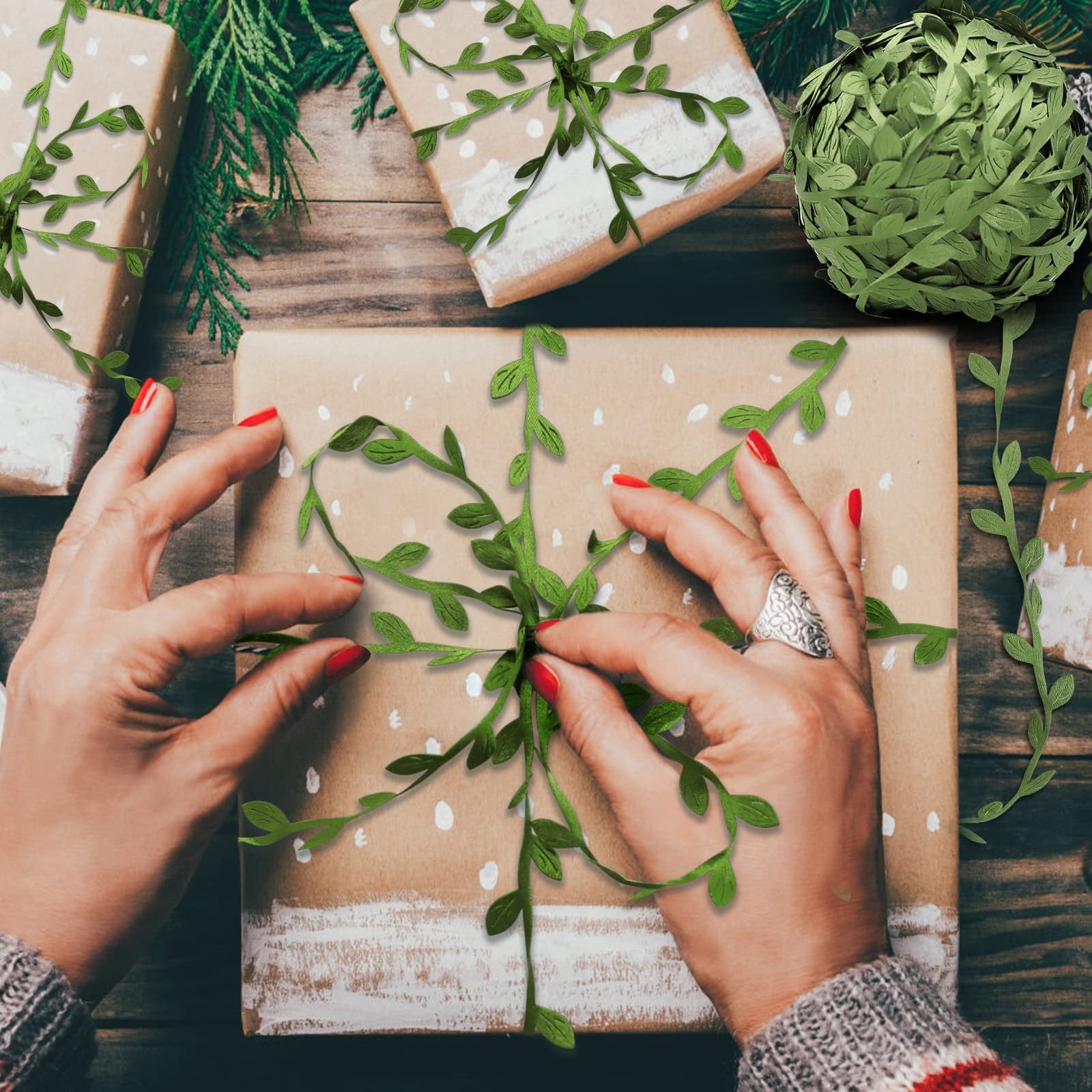 Hands decorating a gift box with artificial leaf vine ribbon, surrounded by other wrapped gifts and greenery.