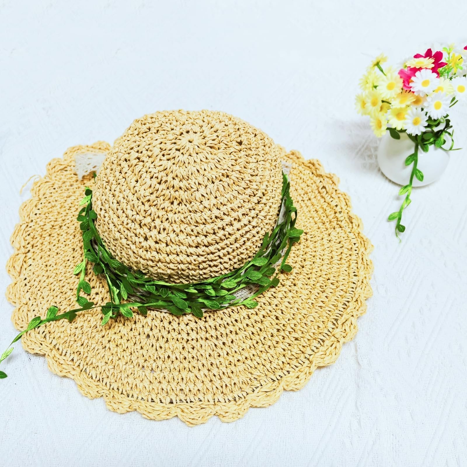 Woven straw hat with green artificial leaf vine draped around the crown, next to a small vase of flowers.
