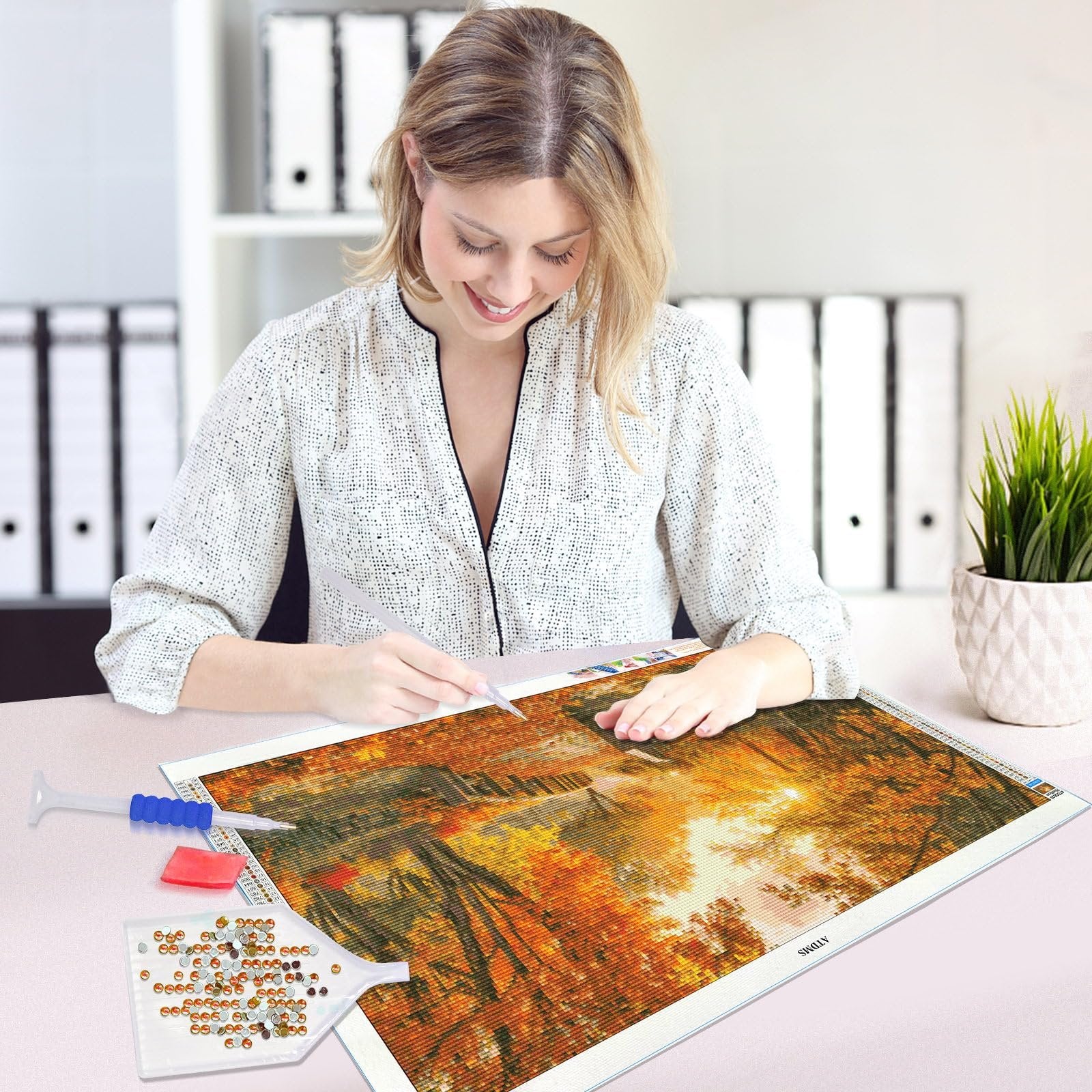 Woman working on a forest sunset diamond painting kit with tools and diamonds laid out on a desk.
