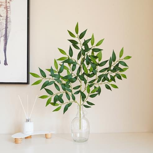 Artificial Italian Ruscus greenery stem in a clear glass vase on a white surface, with a framed picture and diffuser in the background.