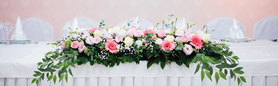Table centerpiece arrangement with pink and white roses, gerbera daisies, and greenery on a white tablecloth.
