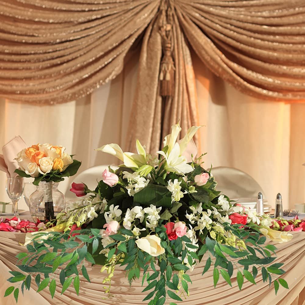 Artificial Italian Ruscus greenery stems draped across a decorated table with floral arrangements and draped curtains in the background.