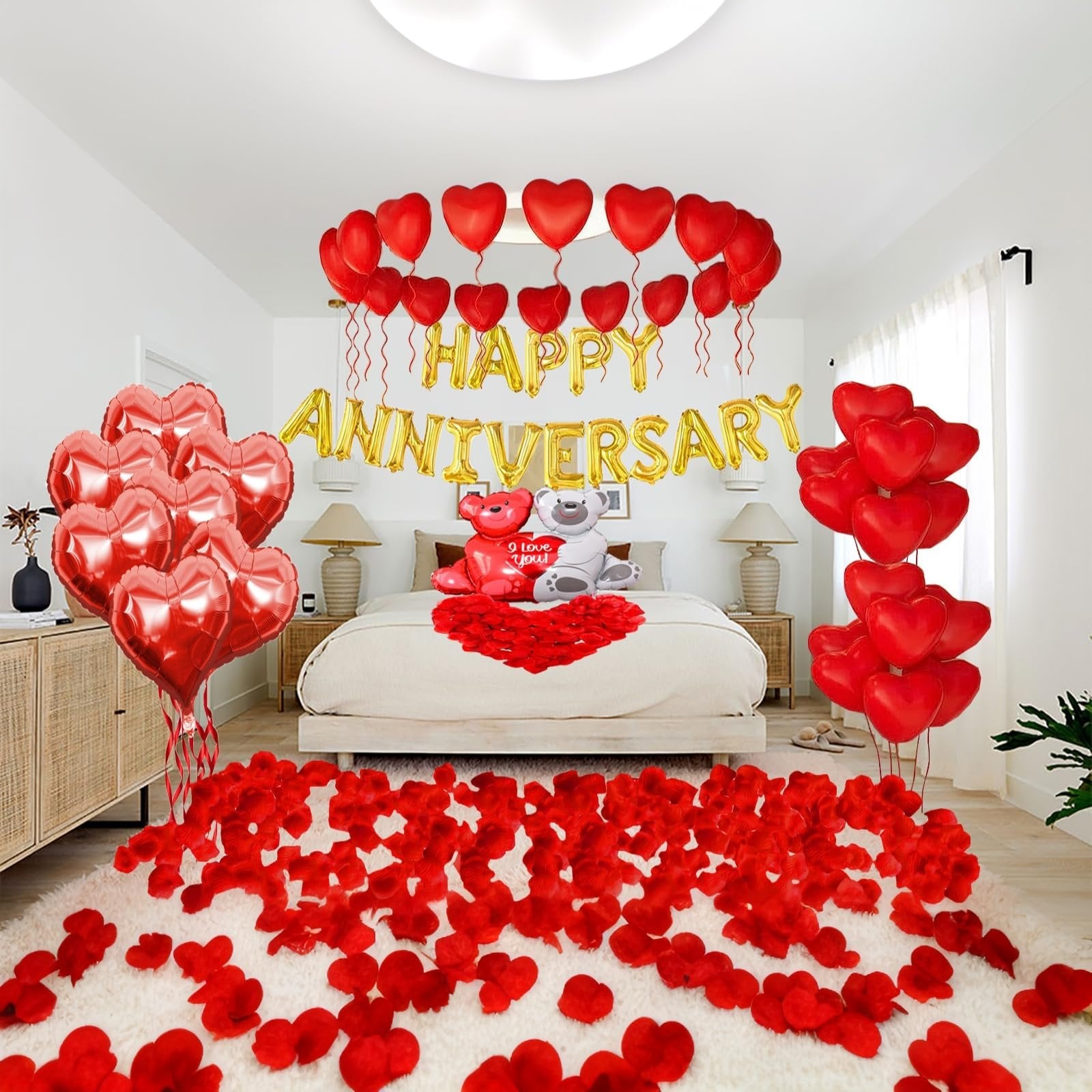 Anniversary bedroom decor with red heart balloons, gold foil 'Happy Anniversary' banner, two teddy bears, and scattered red rose petals on the floor and bed.
