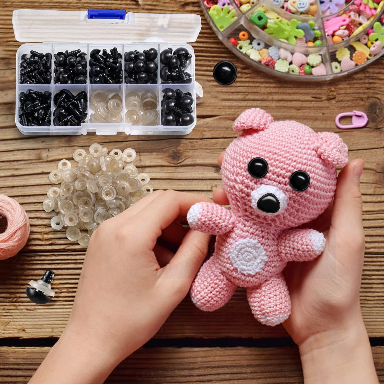 Hands holding a pink crocheted bear with black safety eyes and nose, surrounded by a variety of black safety eyes, noses, and clear washers in a divided plastic case.