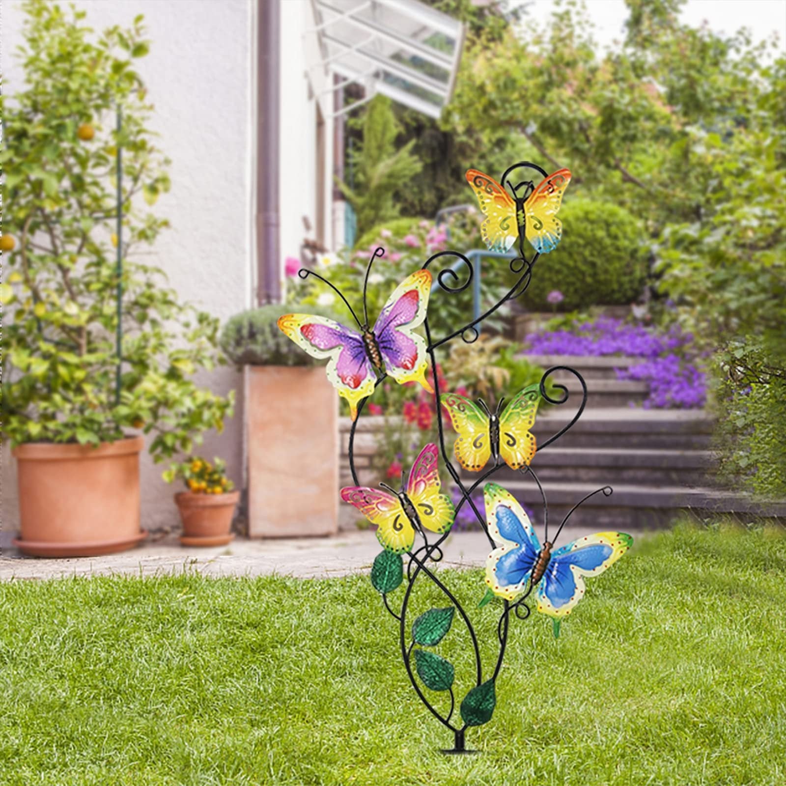 Metal garden stake with five colorful butterflies and green leaves, set in a garden with a house and stairs in the background.