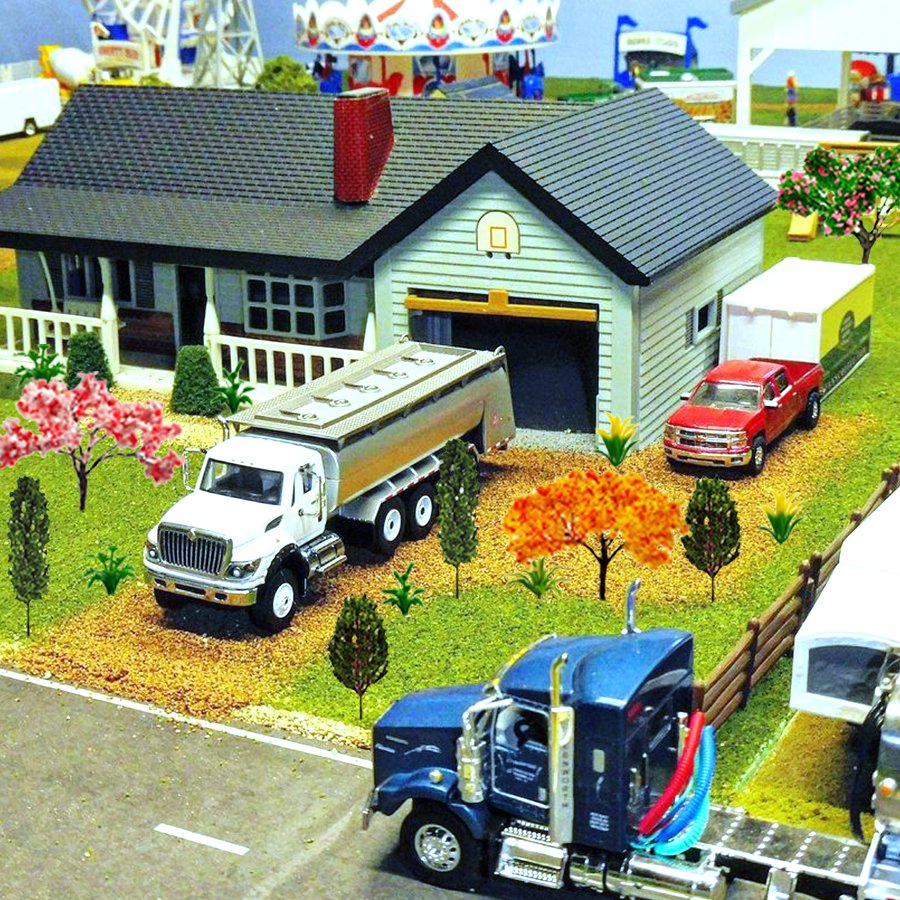 Diorama scene with miniature house, garage, and various trucks on a road and lawn.