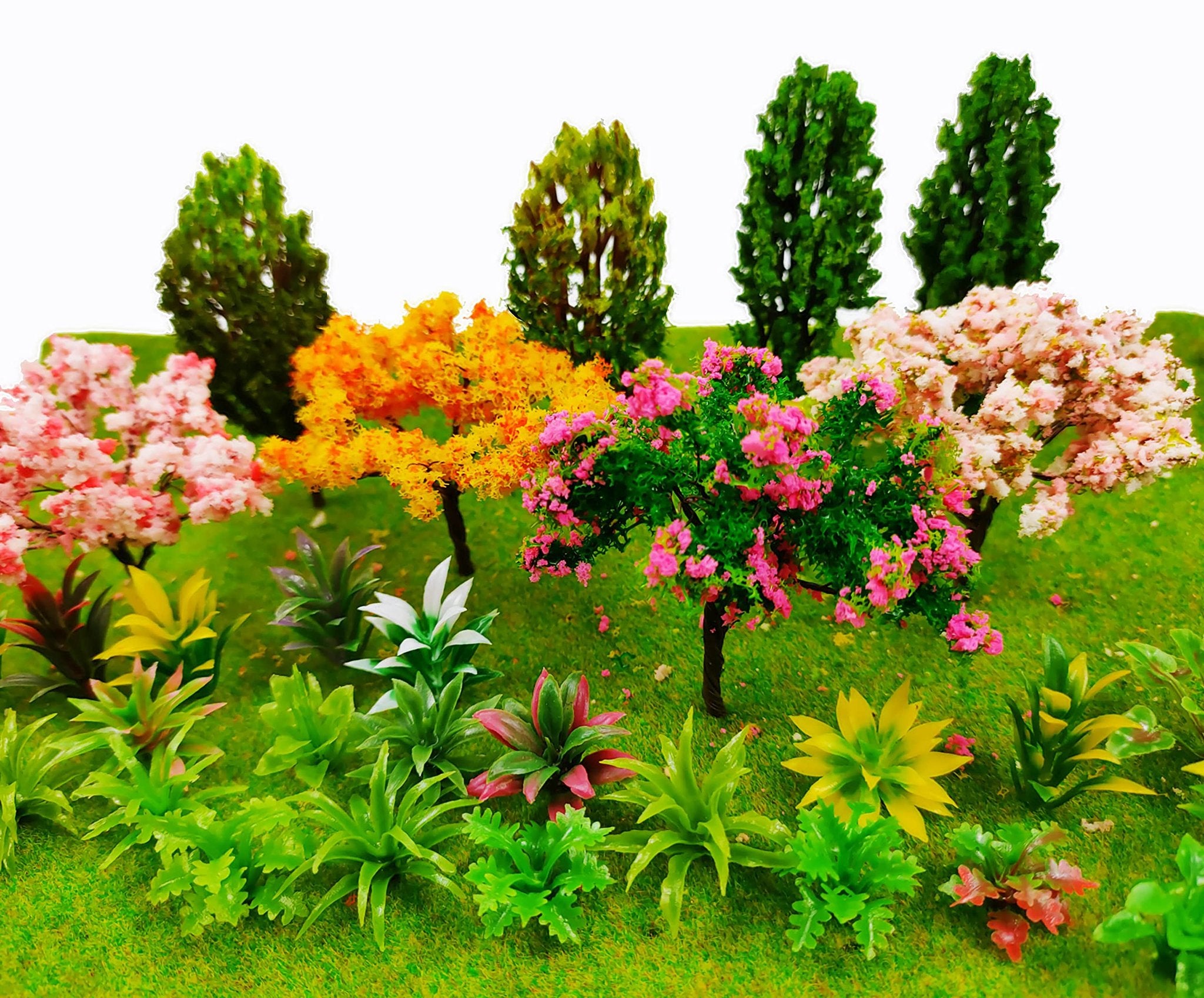 Assortment of miniature trees and plants on green textured ground, featuring pink, yellow, and green foliage.