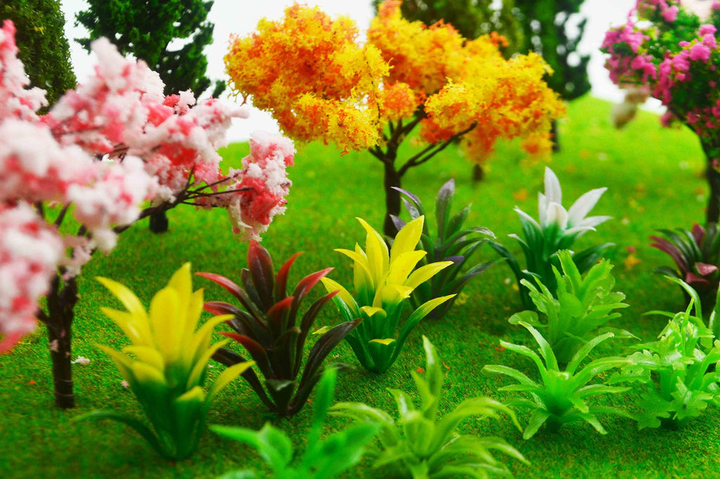 Assortment of miniature model trees and plants in various colors, including pink cherry blossoms, yellow and orange foliage trees, and green and red plants, arranged on a grassy landscape.