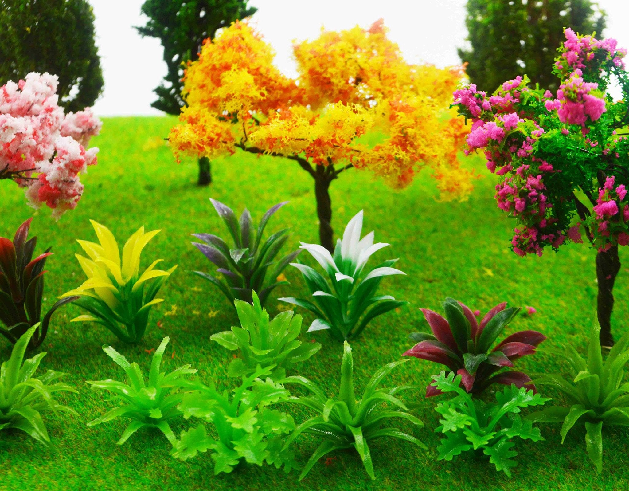 Assortment of miniature artificial trees and plants on green grass, including pink, yellow, and orange foliage.