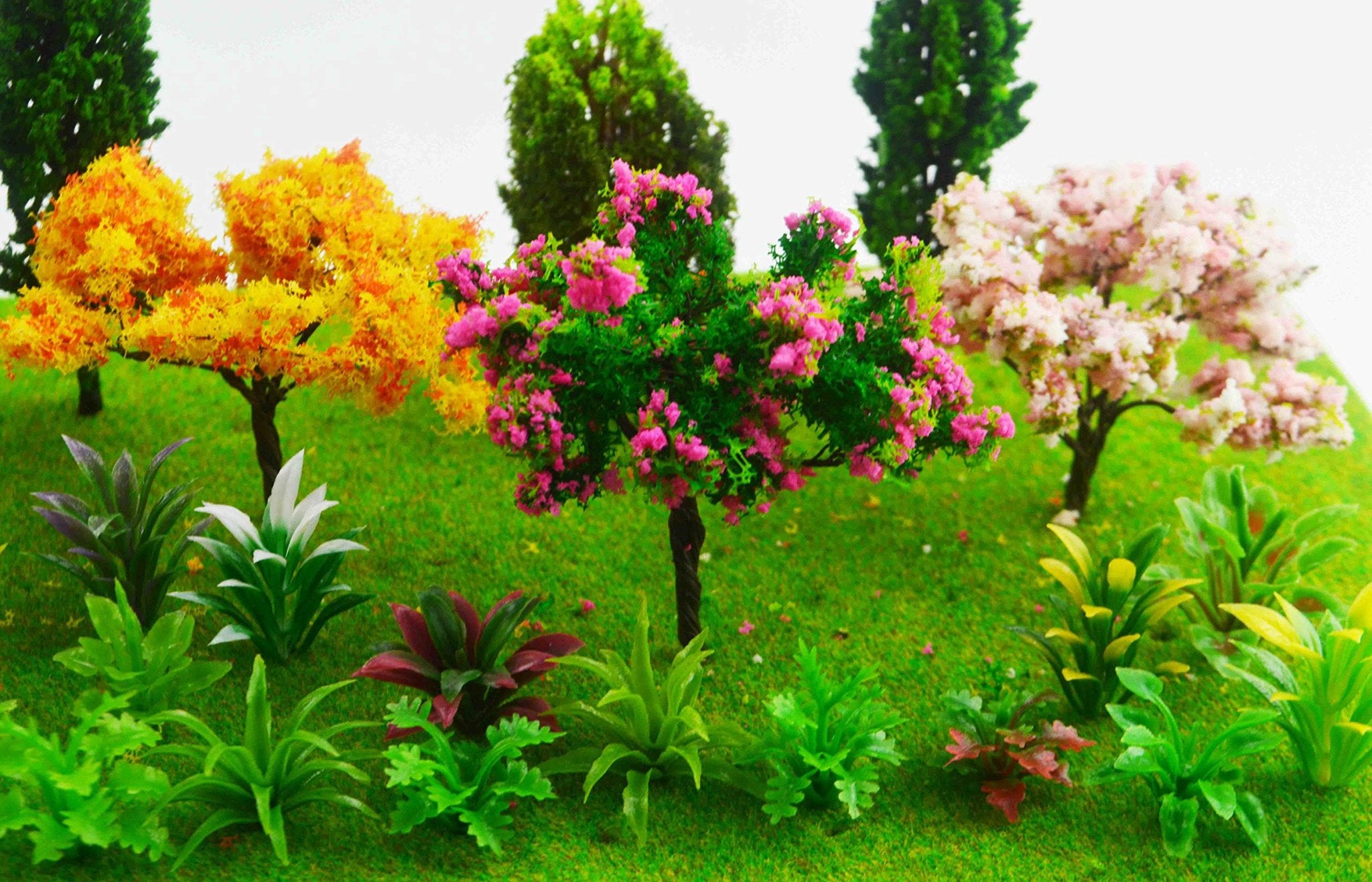 Assortment of miniature artificial trees and plants on a green grassy base, including yellow, pink, and green foliage.