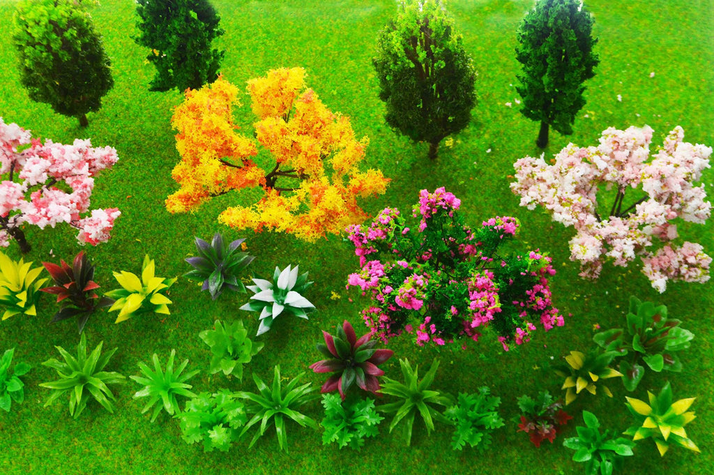 Assortment of miniature trees and plants on green artificial grass, including pink cherry blossom, yellow and orange leafed trees, and various green and red succulents.