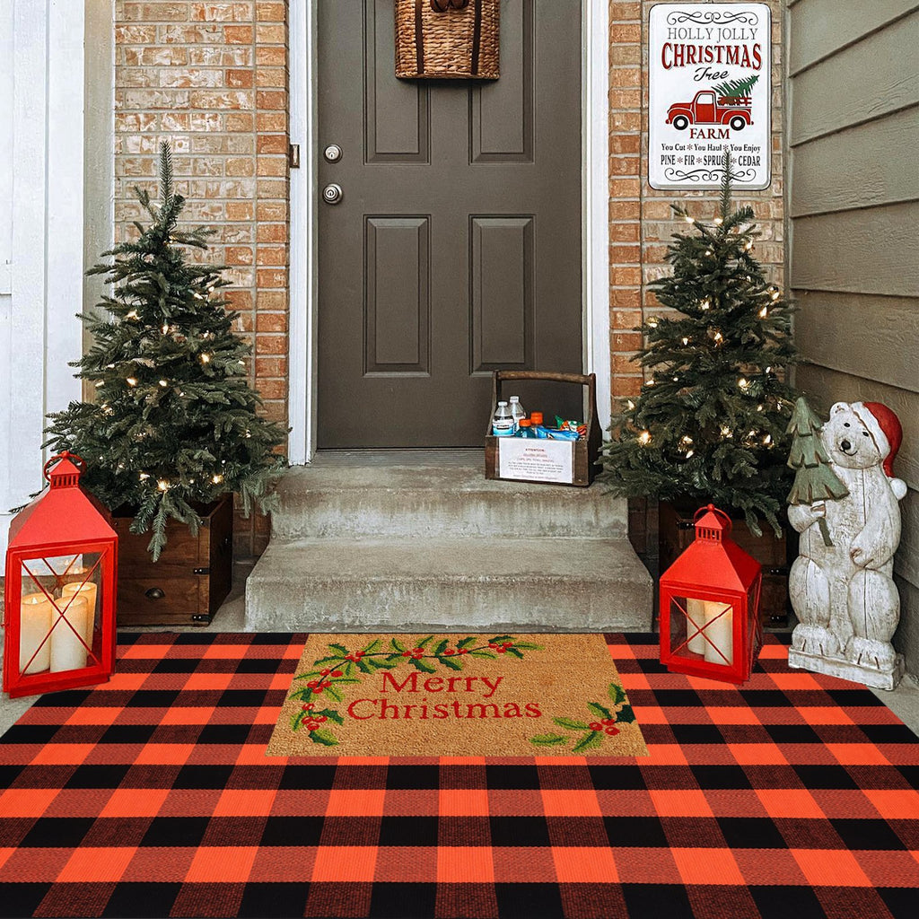 Orange and black buffalo plaid rug layered with a Merry Christmas doormat on a porch with decorated trees and lanterns.