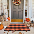 Orange and black buffalo plaid rug on a porch with fall decorations including pumpkins and hay bales.