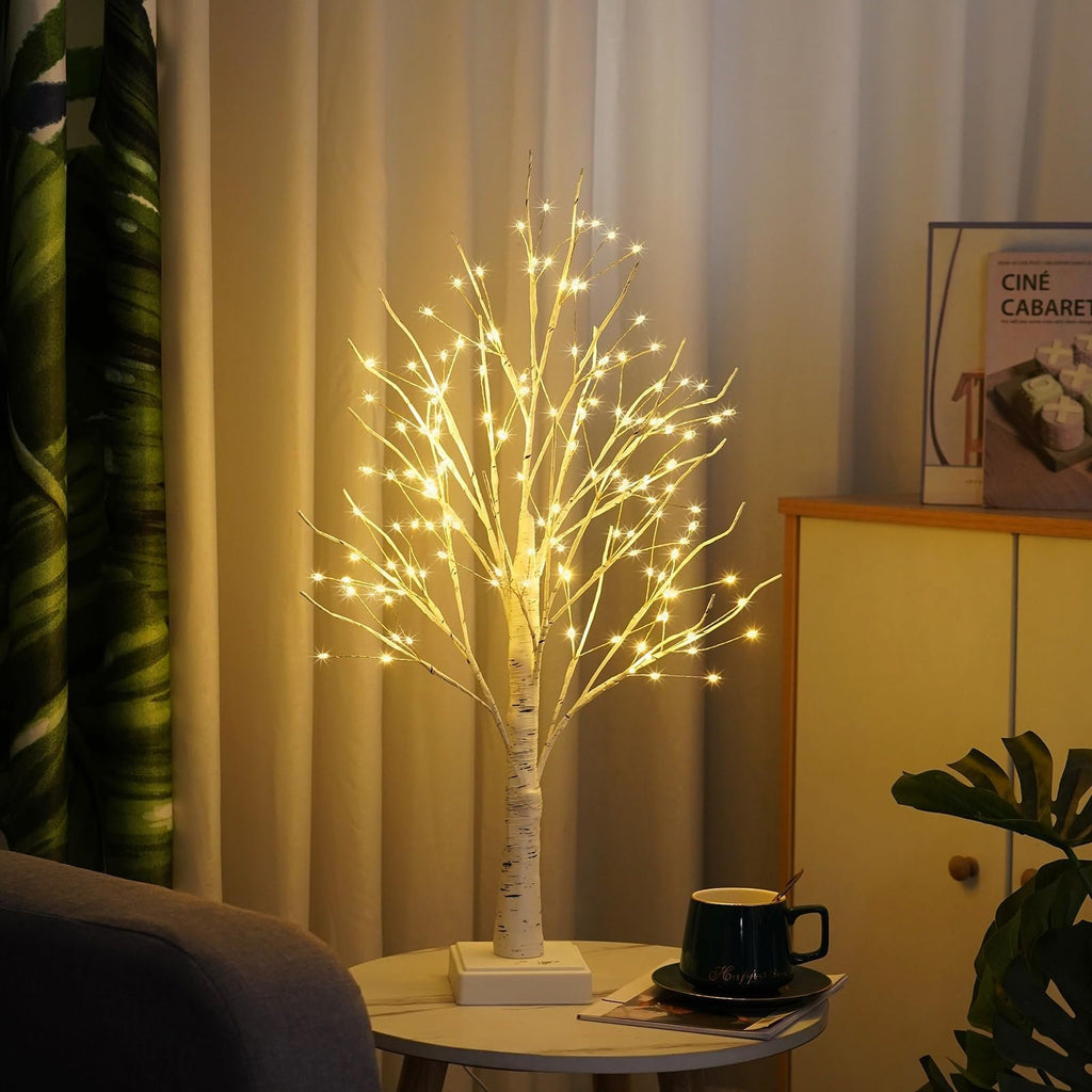 White birch tree lamp with warm white LED lights on a side table with a book and mug.