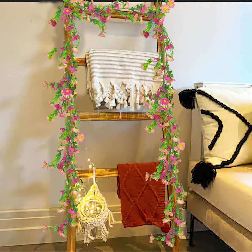 Artificial silk daisy vine garland with pink flowers and green leaves draped on a wooden ladder with blankets and lights.