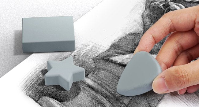 Three grey art erasers in different shapes (rectangle, star, triangle) on a sketchpad with pencil drawings. A hand holds the triangle eraser.