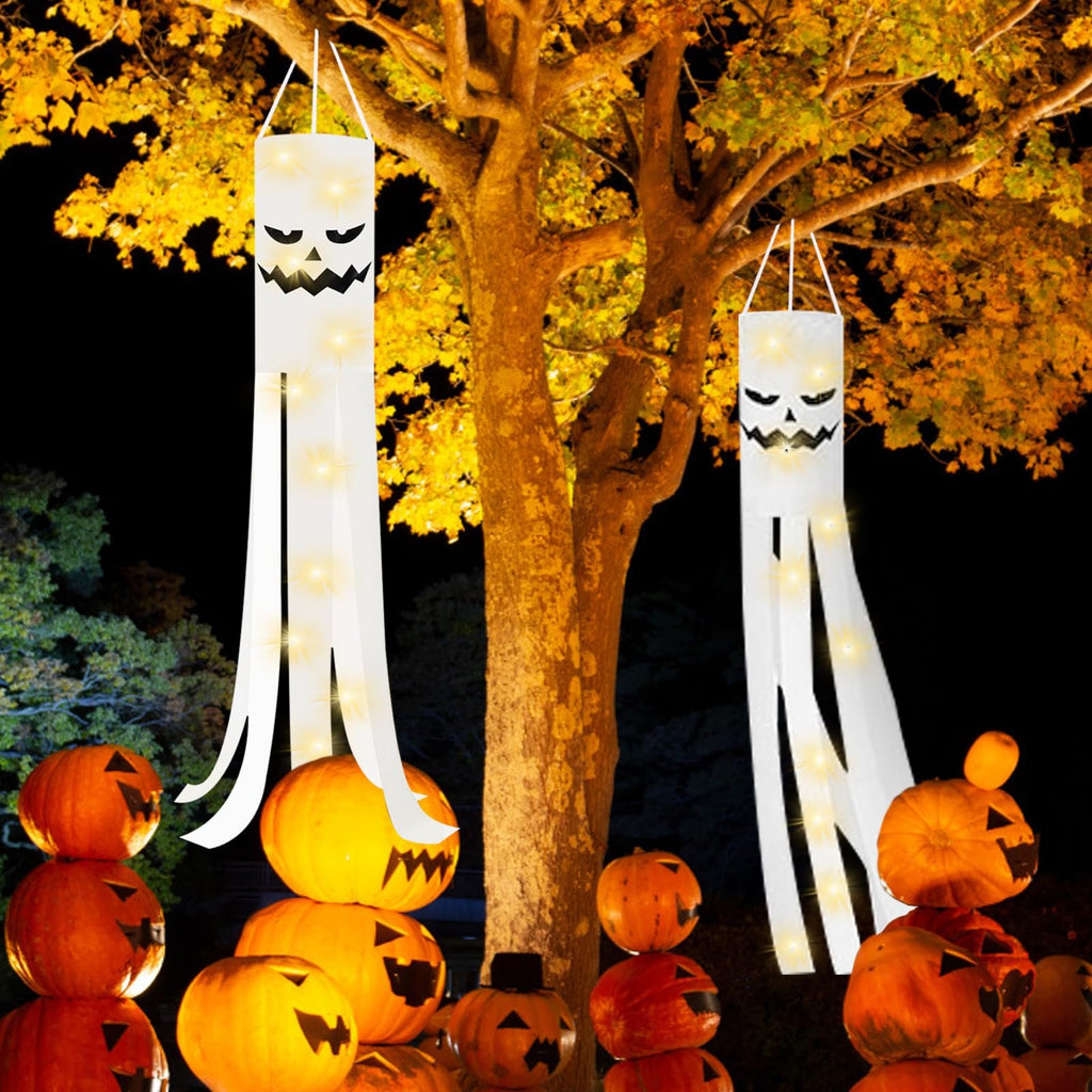 Two white Halloween ghost windsocks with jack-o-lantern faces and string lights hang from a tree, surrounded by carved pumpkins.